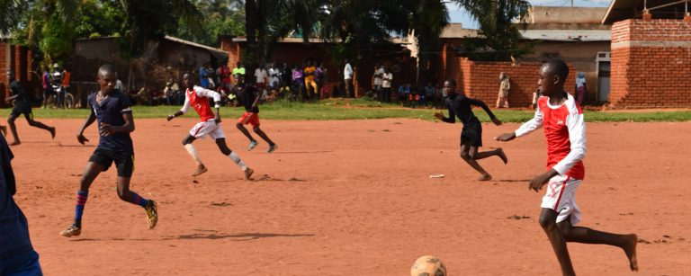 Boys playing a football match on a red dirt pitch