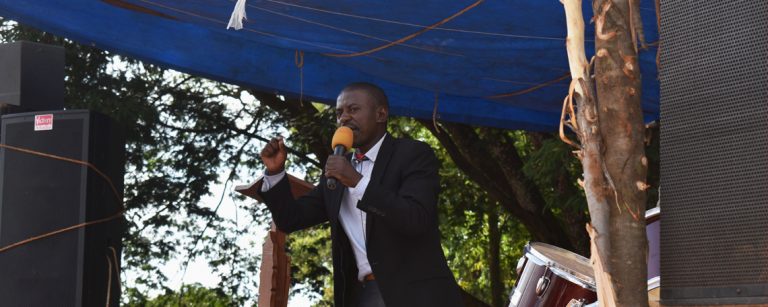 a man preaches on a stage with a drum kit and large speakers. A crowd watches from all sides