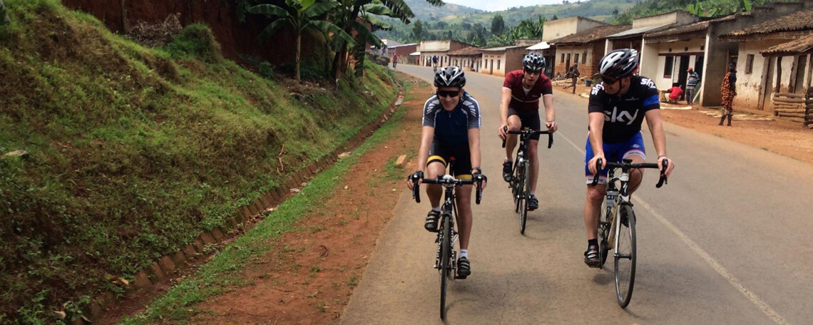 three men on bikes smile as they cycle along a road, a village to the right and a green slope to the left