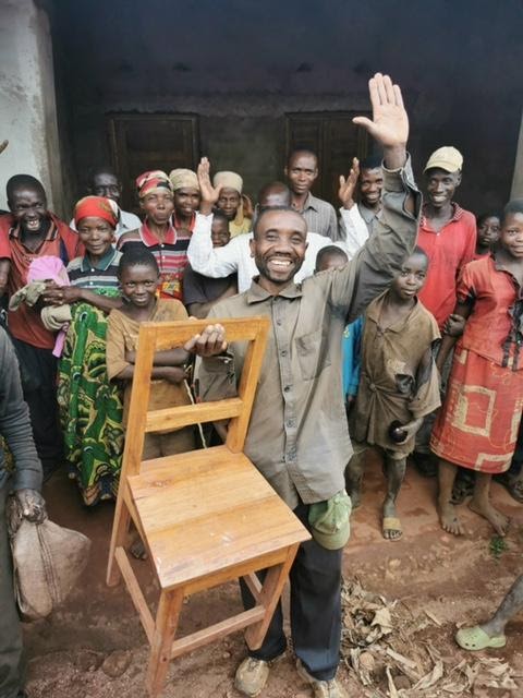 Etienne holds up a chair he has made and smiles, surrounded by his community